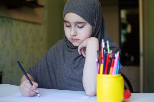flicka i 11-12 års åldern ikläddi grå hijab som sitter vid ett skrivbord och ritar
