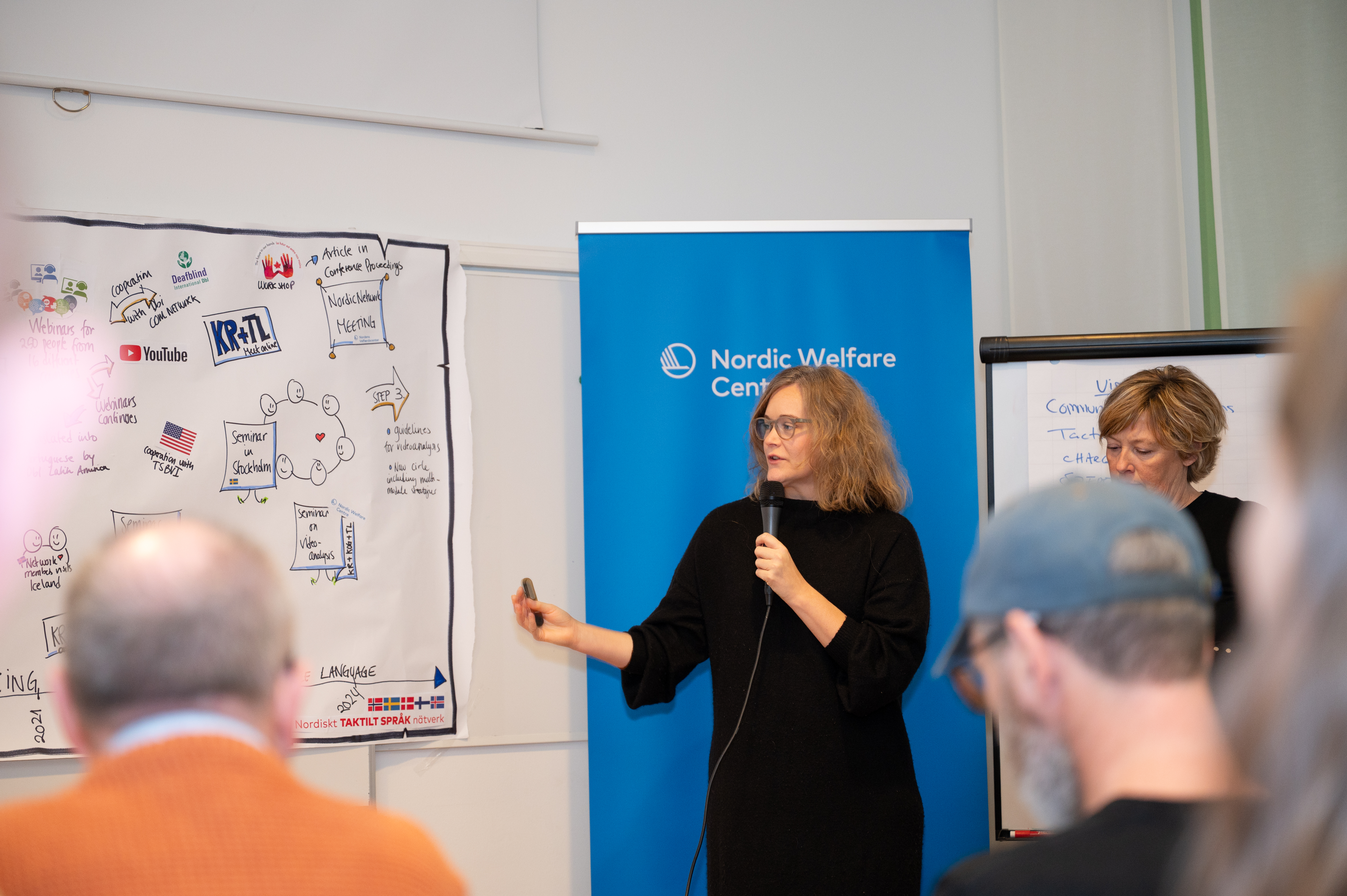Woman presenting with a microphone in front of a Nordic Welfare Centre banner and illustrated poster.