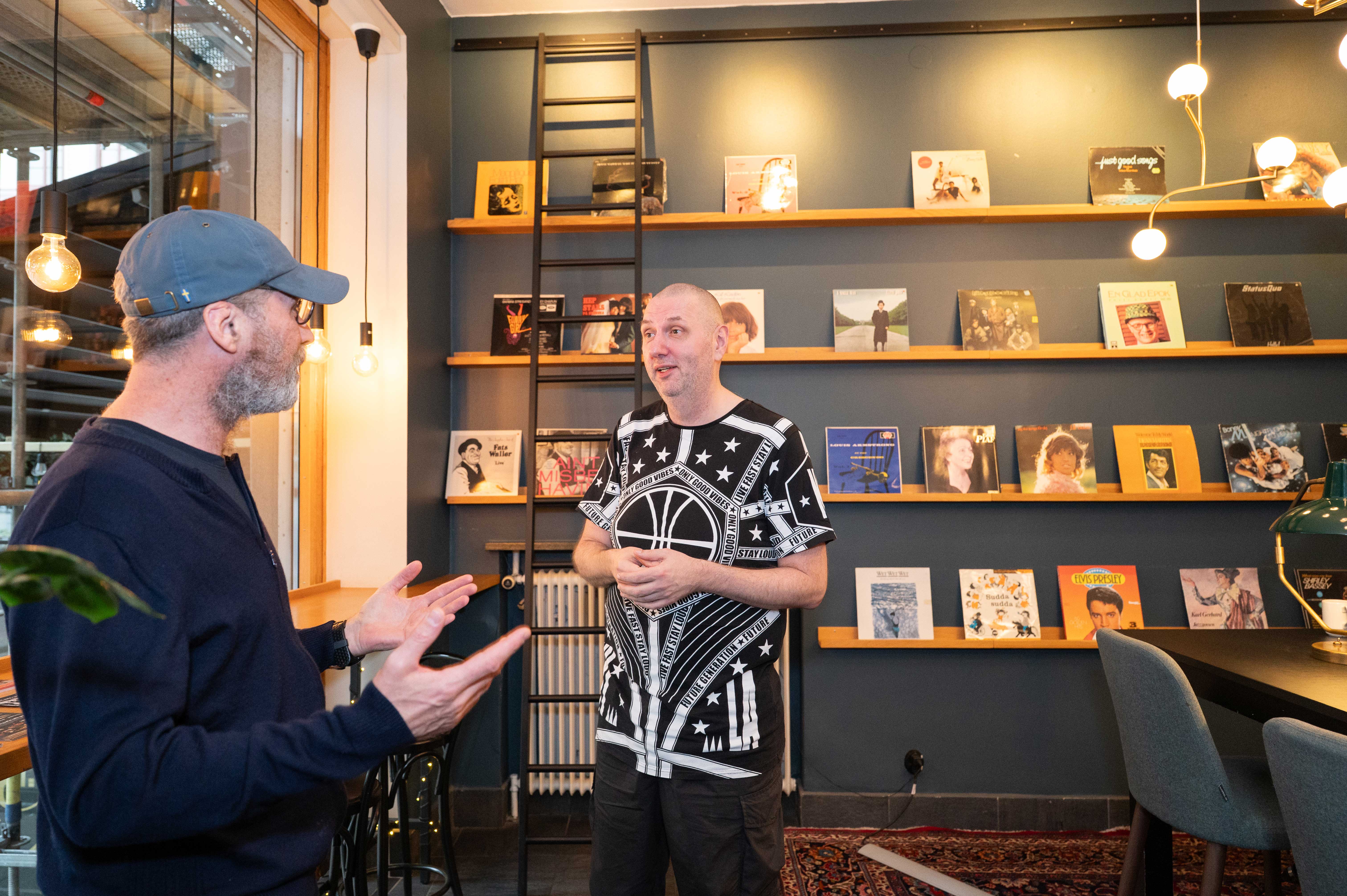 Two people having a conversation in a cozy room with books and records on display shelves.