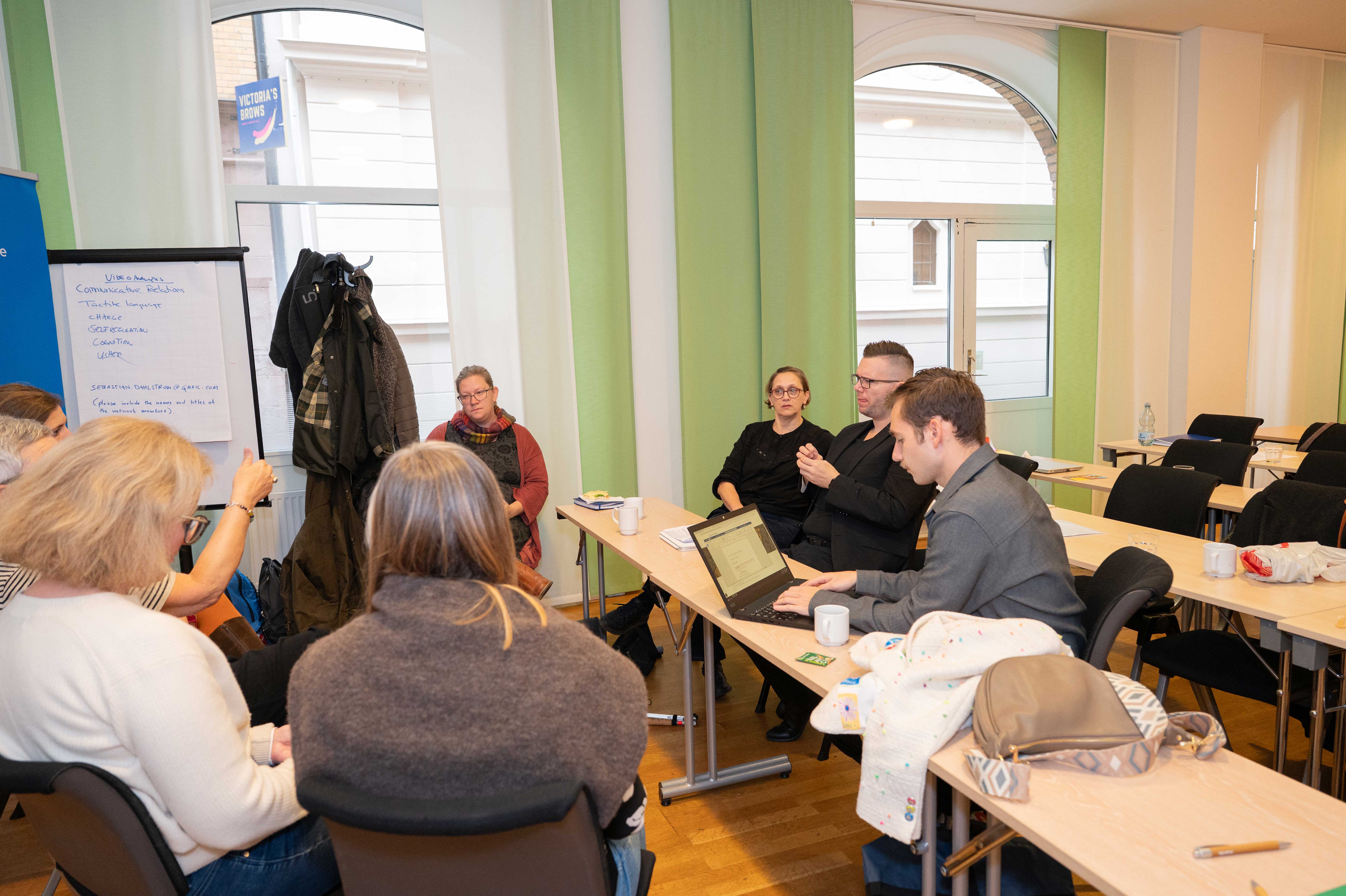 Group of people sitting in a classroom setting, engaged in discussion with a whiteboard in the background.