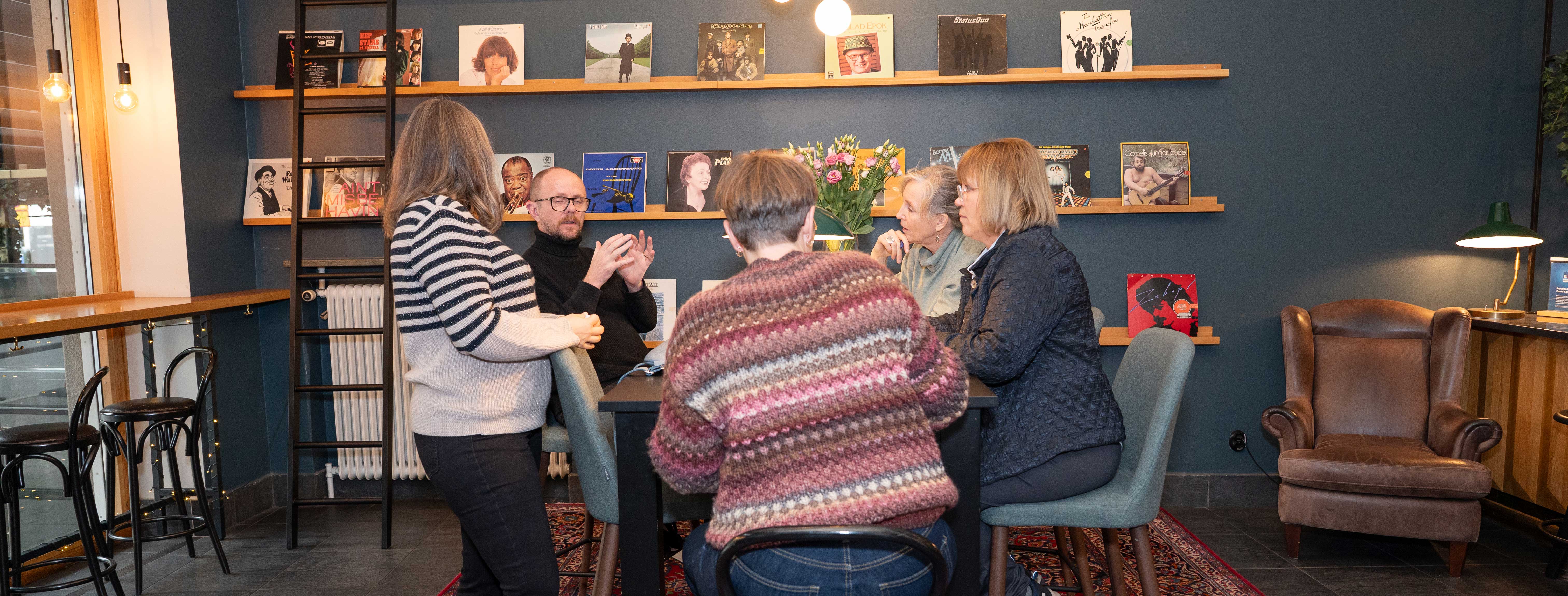 Group of people gathered around a table in a cozy room with books and records on display, engaged in conversation.