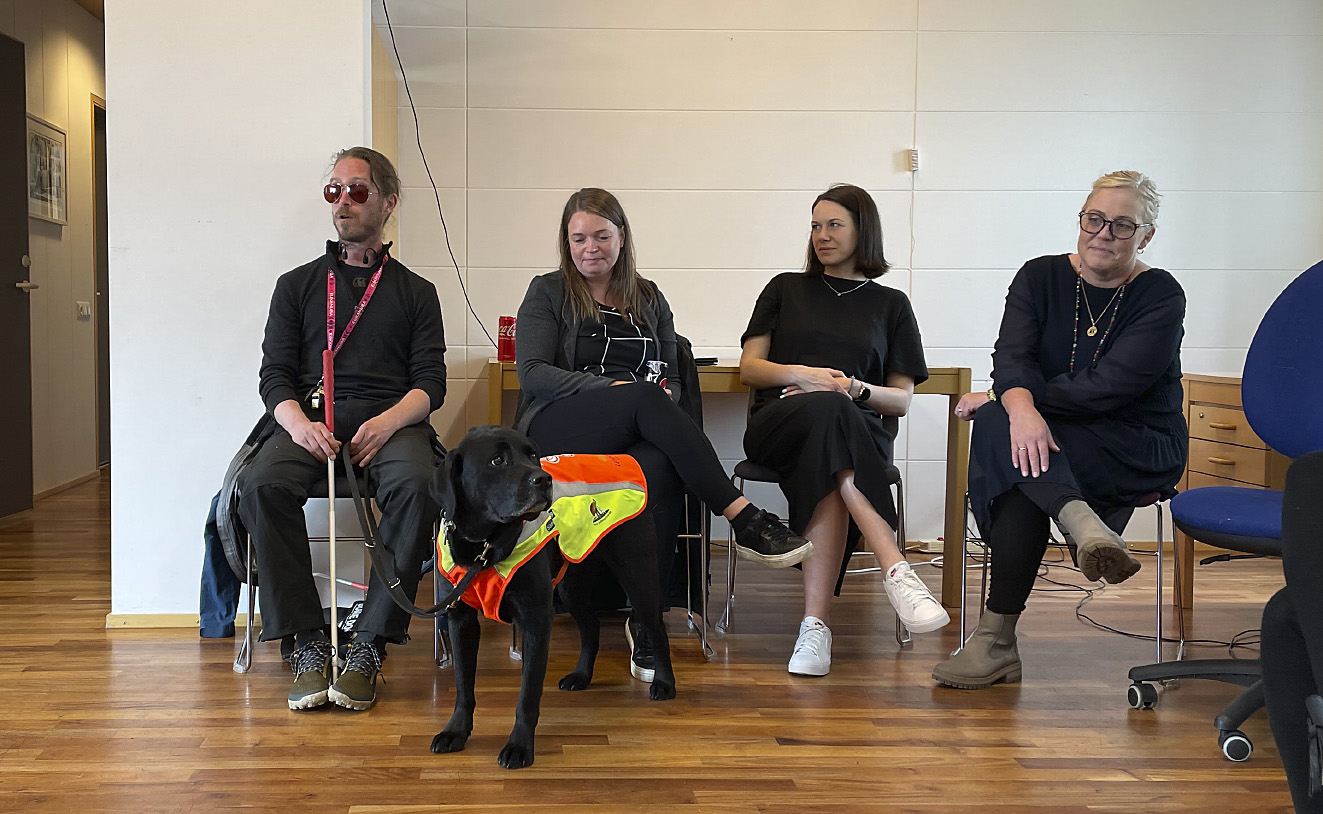 Et bilde av en assistenthund med refleksvest stående foran en gruppe med sittende personer. Innendørs.