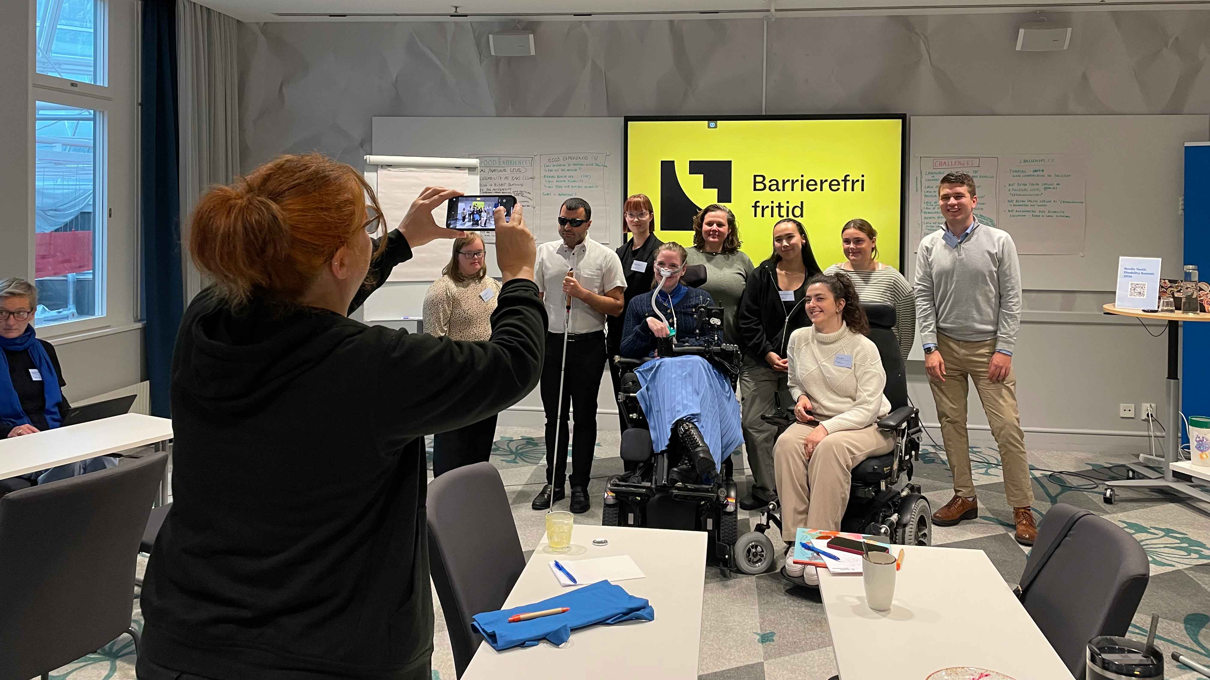 9 ungdommer står i et konferanserom og blir fotografert av en person som er uskarp i forgrunnen. 
