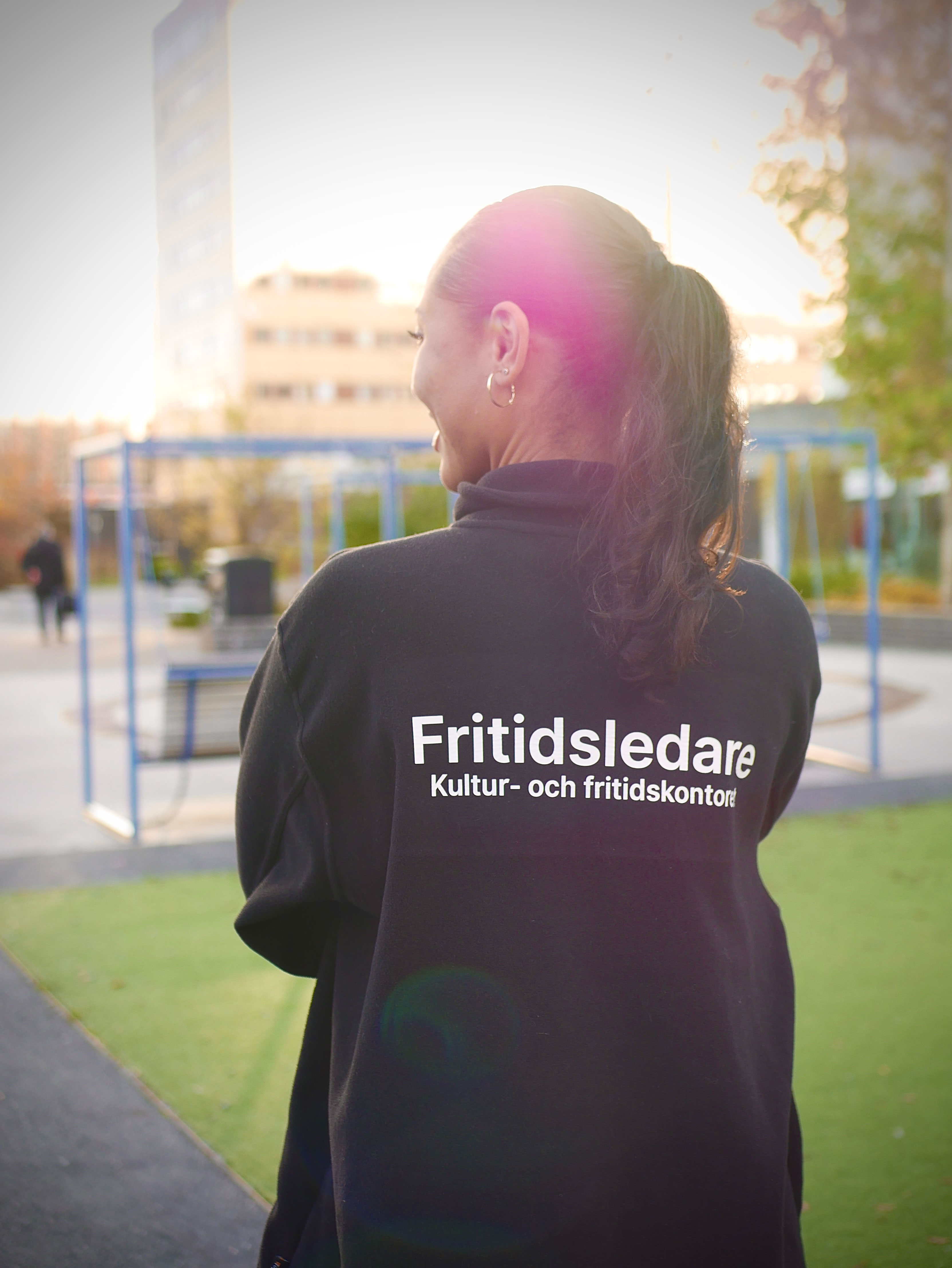 A youth worker wearing a black jacket with the text ‘Fritidsledare – Kultur- och fritidskontoret’ stands outdoors in a playground with sunlight in the background.