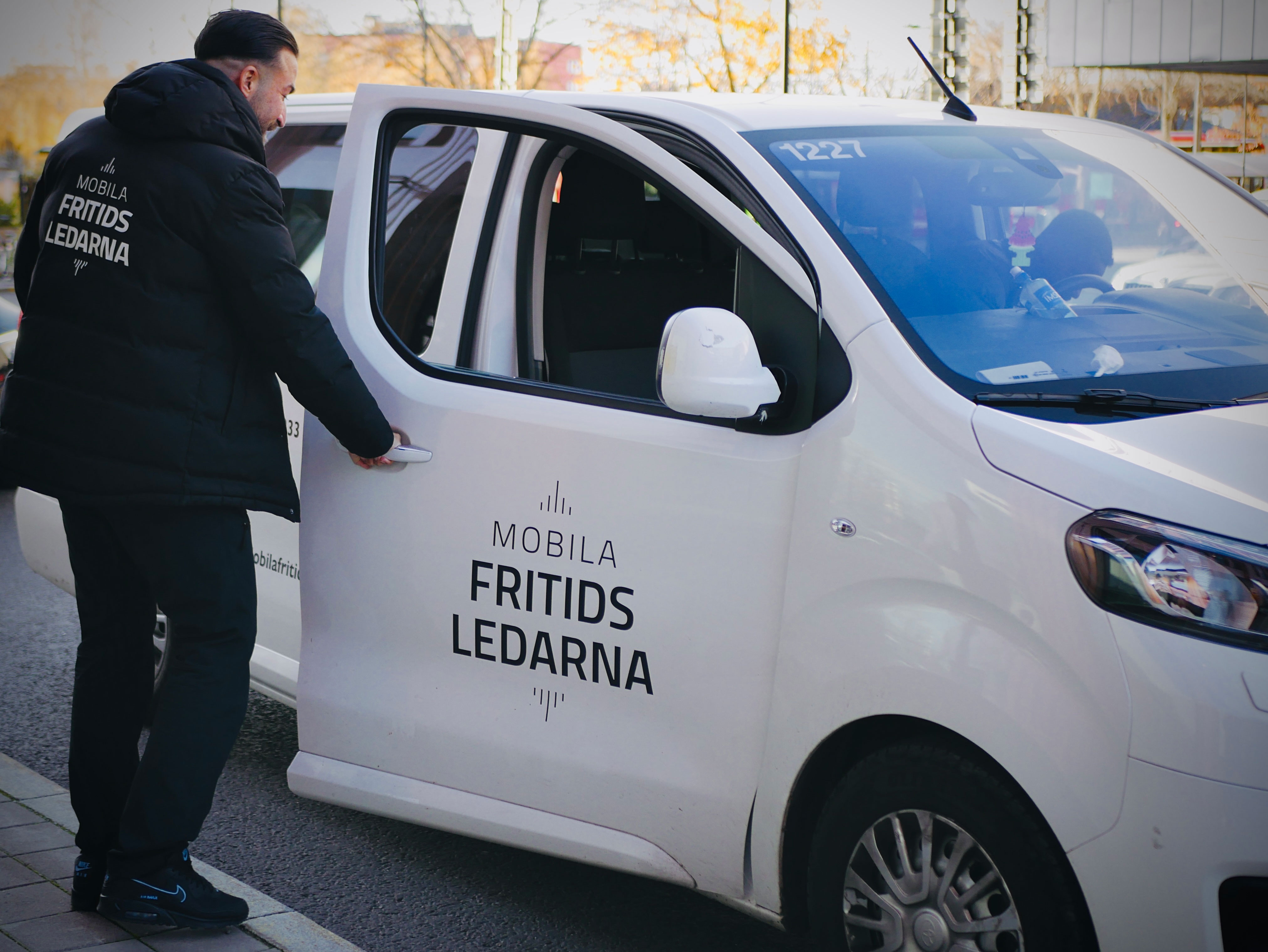 A man wearing a jacket labeled ‘Mobila Fritidsledarna’ opens the door of a white van with the same text printed on the side.
