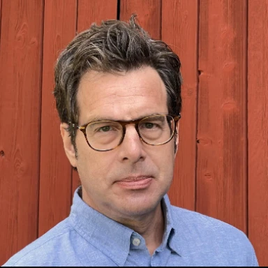 Portrait of Kenneth Nelson, a man with dark hair and glasses, wearing a light blue shirt. He is standing in front of a red wooden wall.
