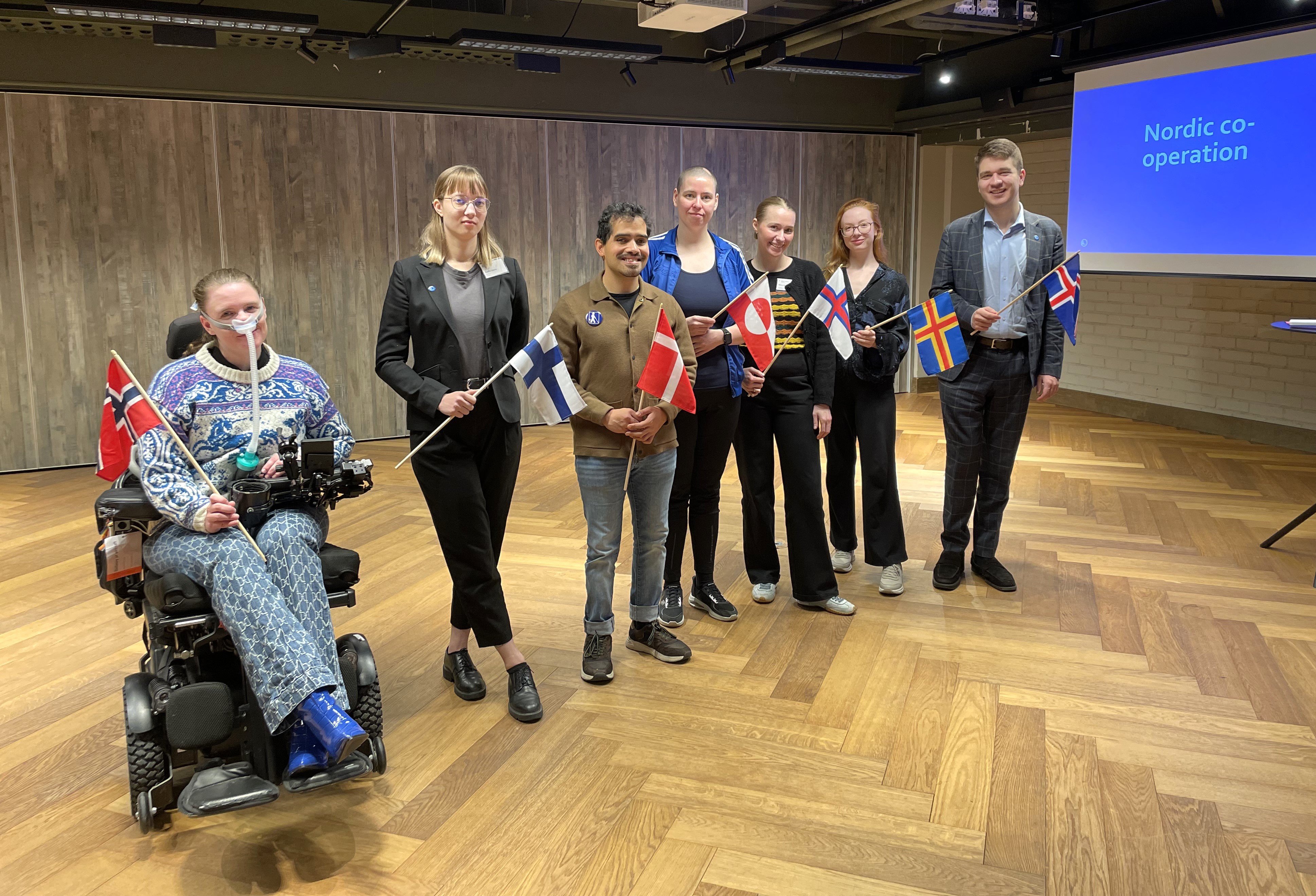 Syv personer som holder flaggene til sine nordiske land står eller sitter i rullestoler i et stort rom.