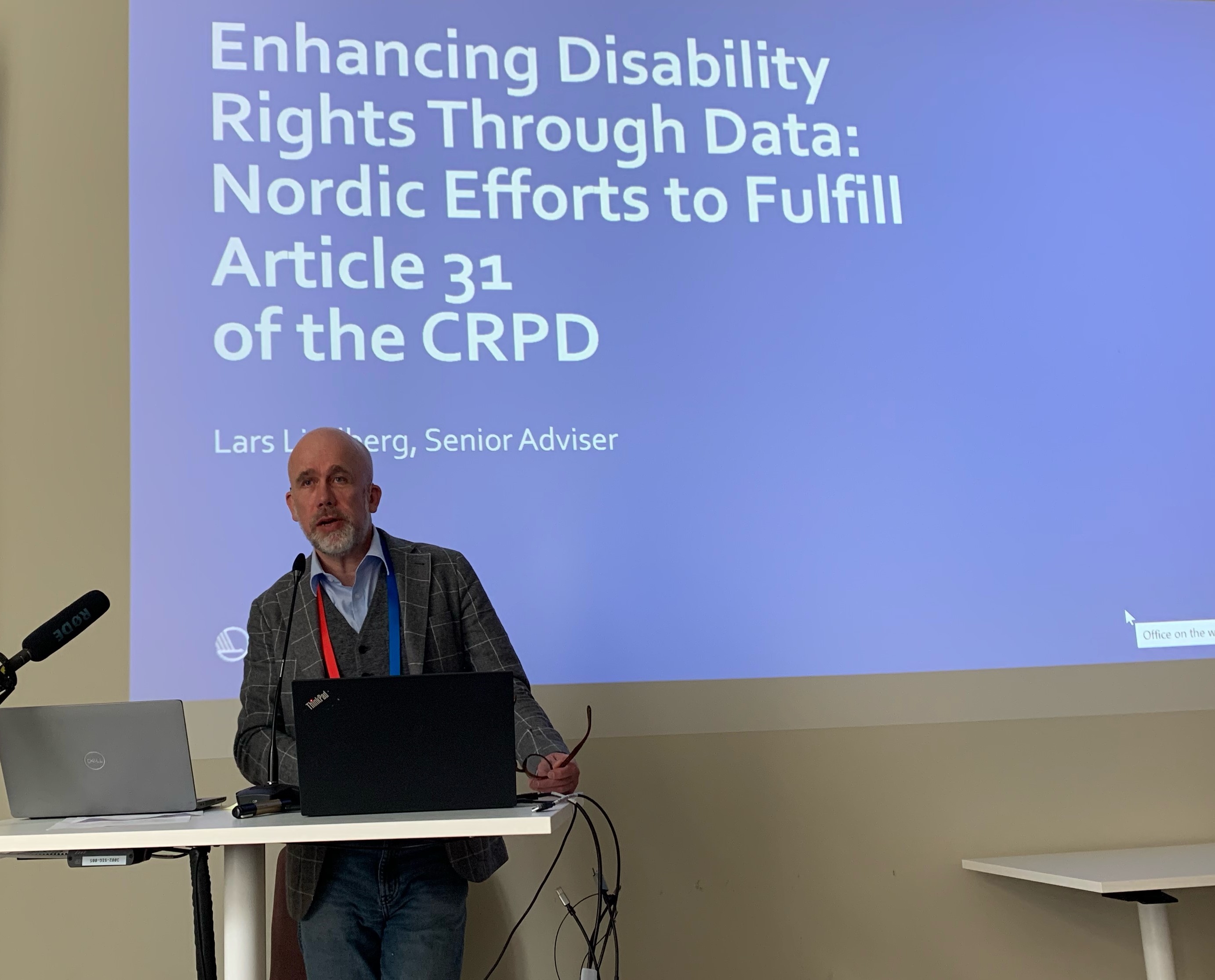 Mann som taler på en konferanse. Tekst bak ham: Styrke funksjonshemmedes rettigheter gjennom data, nordisk innsats for å oppfylle artikkel 31 i CRPD 