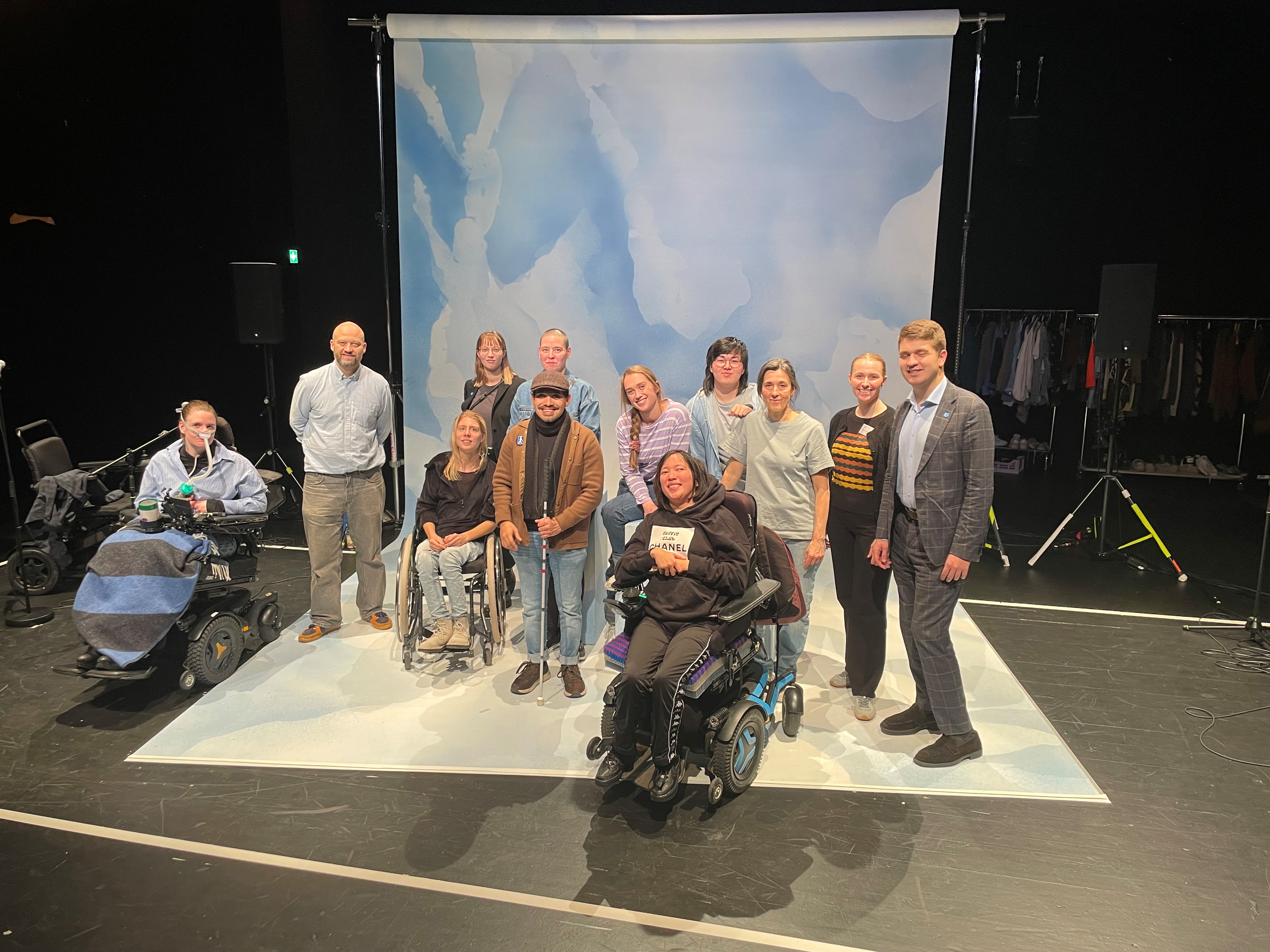 Gruppebilde med 12 personer på en teaterscene med rekvisitter i bakgrunnen. Tre av personene sitter i rullestol, de andre står eller sitter på rekvisitter.