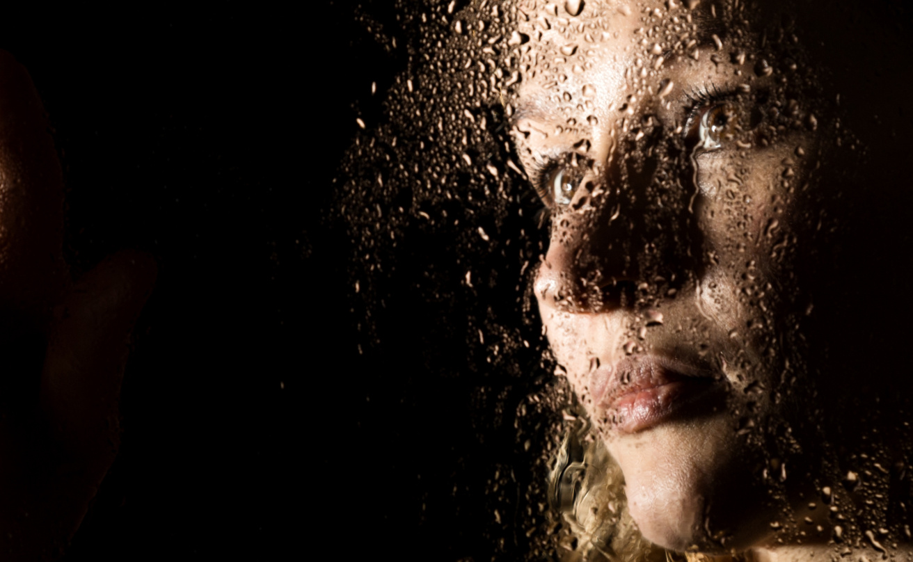 Smooth portrait of sexy model, posing behind transparent glass covered by water drops. young melancholy and sad woman portrait