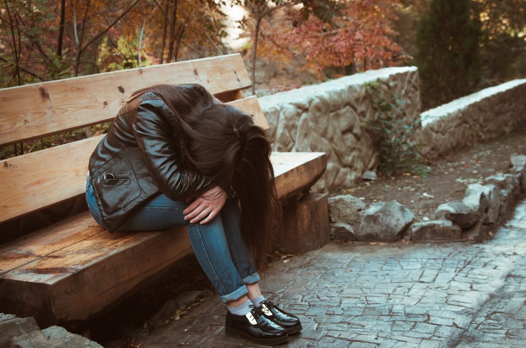 En flicka gömmer huvudet i knät. Hon sitter på en parkbänk.