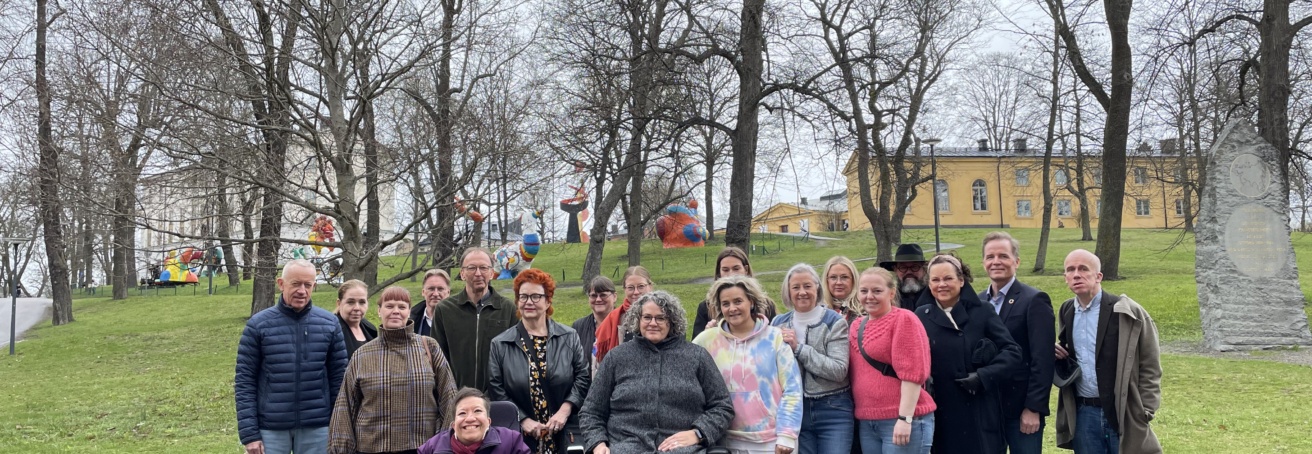 19 personer utomhus, gruppbild, två av dem sitter i rullstol. Grön gräsmatta i bakgrunden.