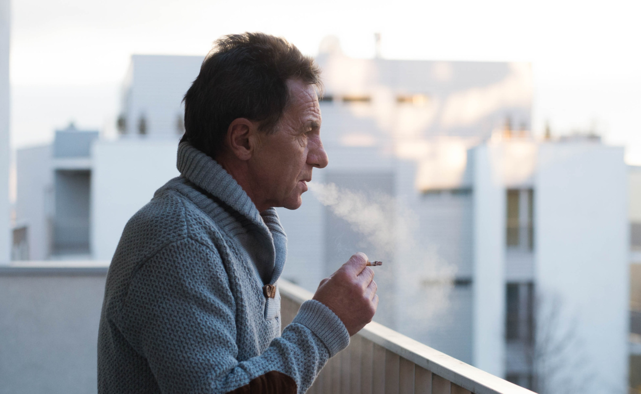 Serious senior man standing on balcony, smoking a cigarette