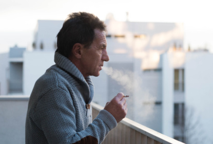 Serious senior man standing on balcony, smoking a cigarette