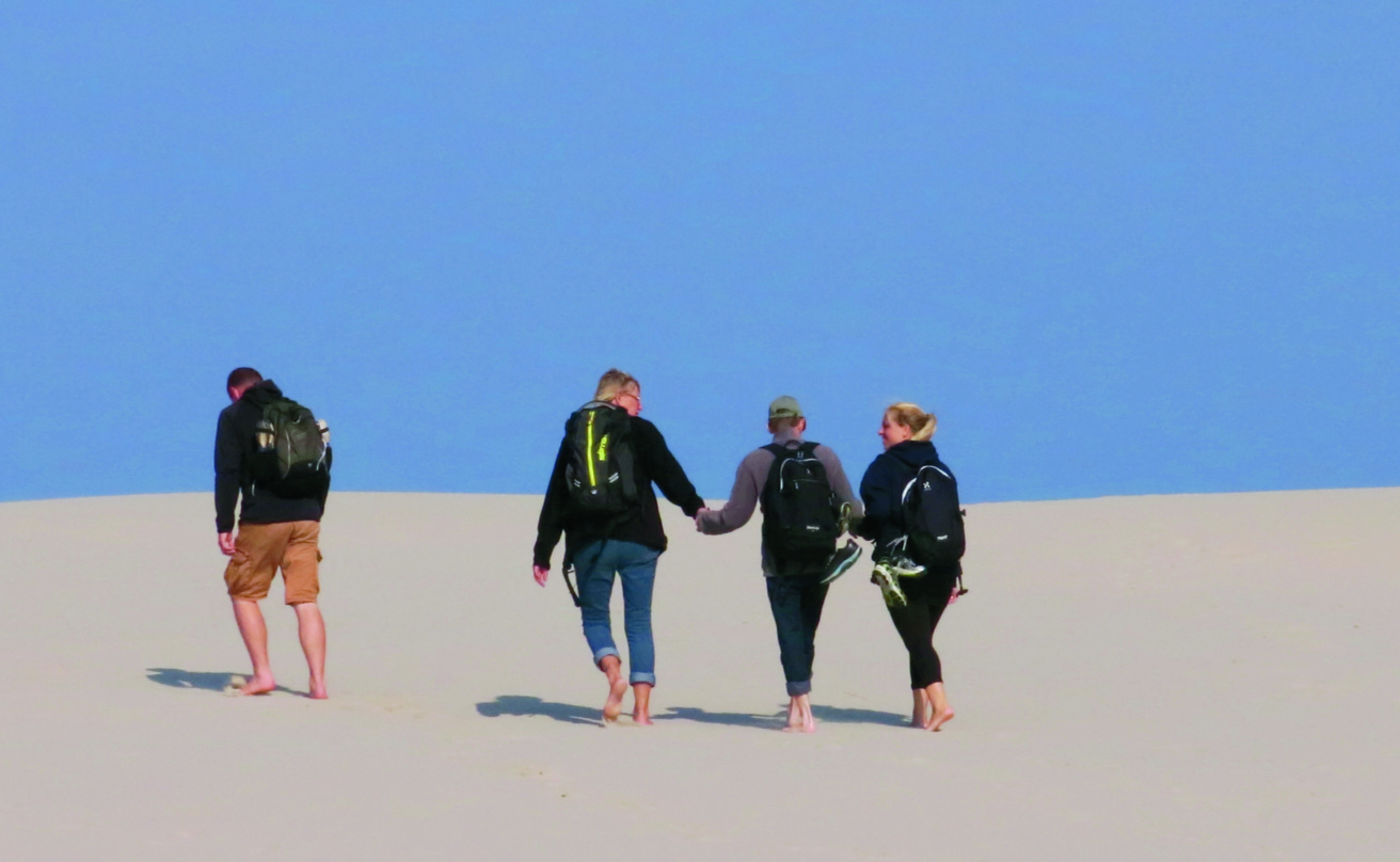 Four people walk barefoot in white sand. Three of them hold hans.