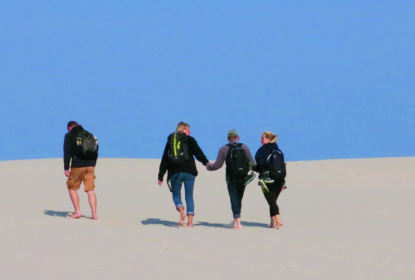 Four people walk barefoot in white sand. Three of them hold hans.