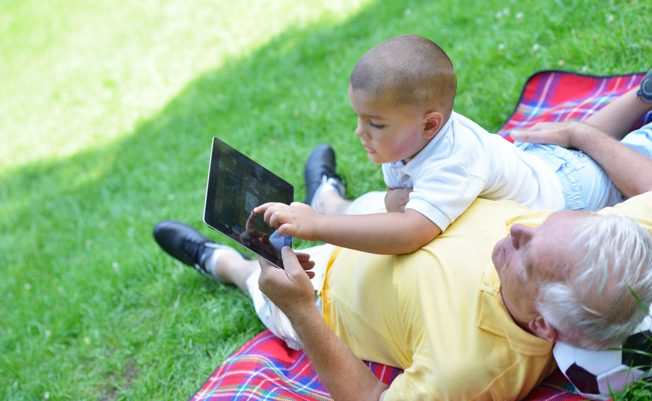 Grandfather and child looking at an ipad while both lying on green grass