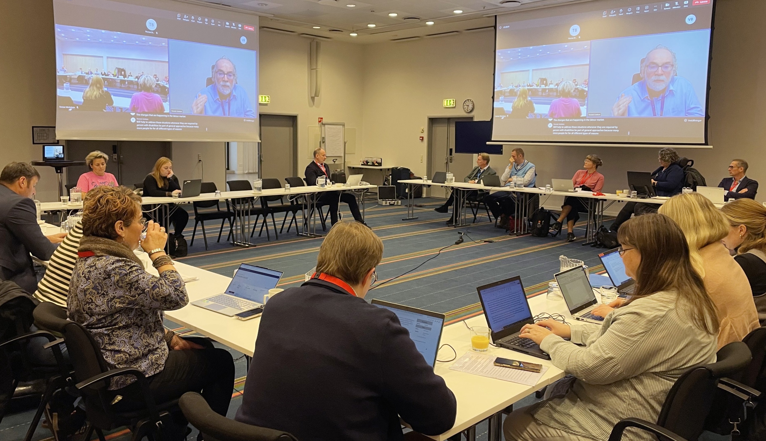 People in a conference room, man giving a speech shown on two screens