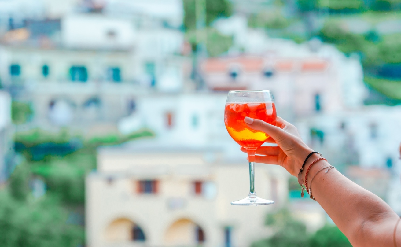 En kvinna håller upp ett glas med Aperol Spritz mot en vacker bakgrund, hus och grönska.