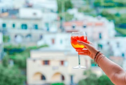 En kvinna håller upp ett glas med Aperol Spritz mot en vacker bakgrund, hus och grönska.