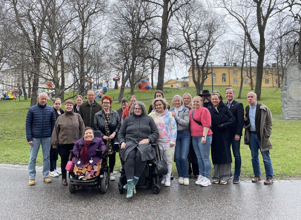 Gruppbild där 19 perosner står utomhus på Skeppsholmen i Stockholm. Två personer är rullstolsburna.