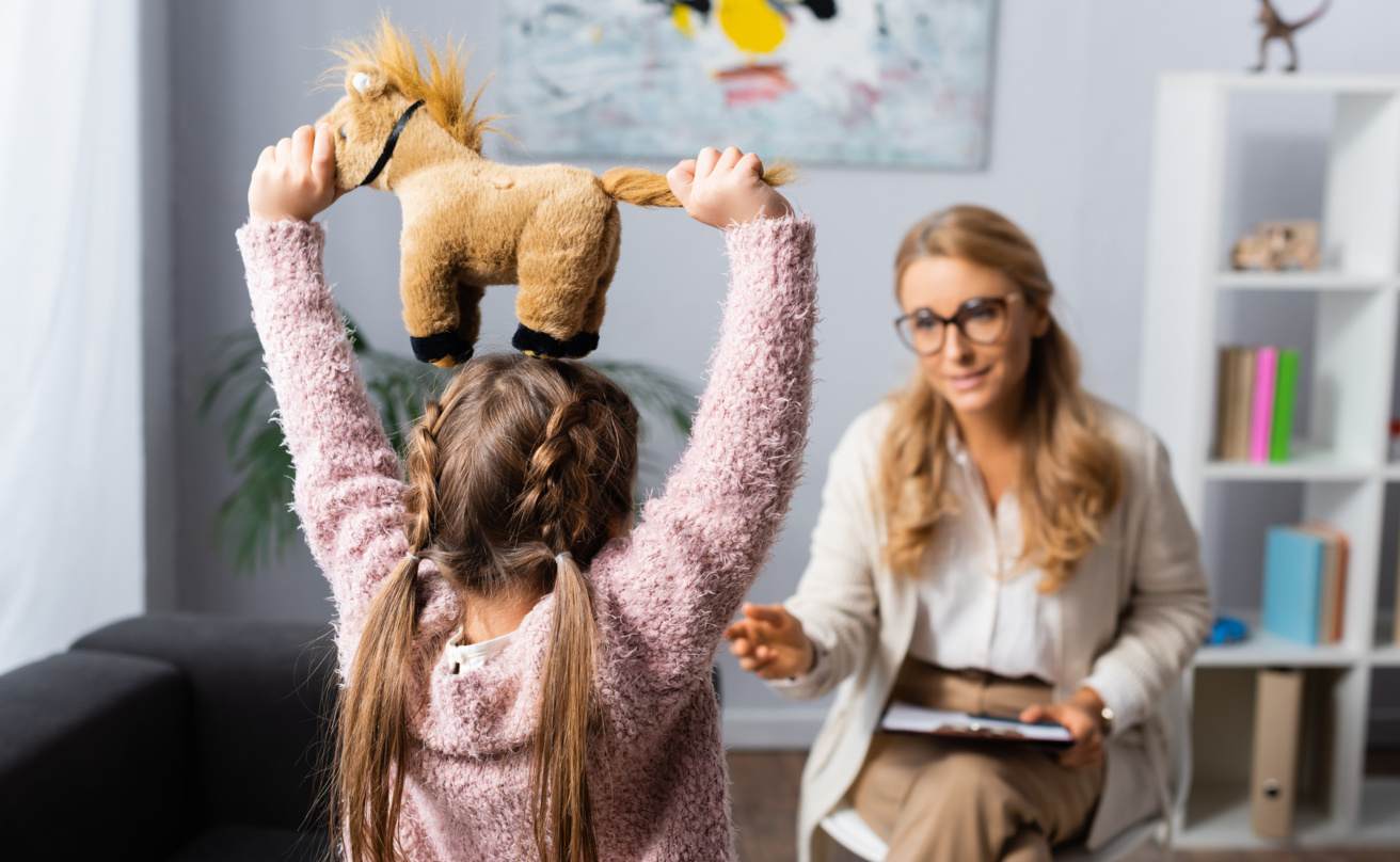 Little girl seen from behind holding a toy above her head. Talking to a woman with clipboard in hand
