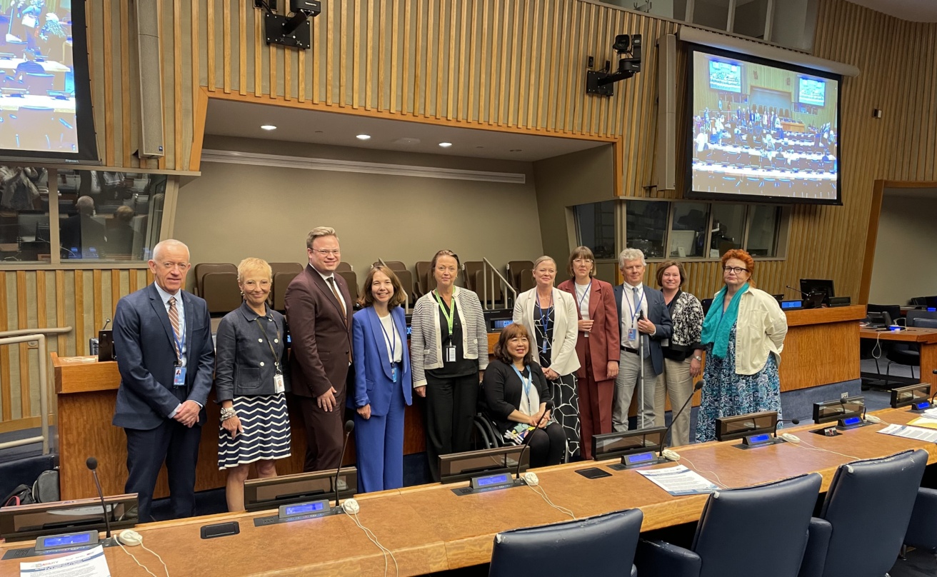 11 people in a conference room at the Nordic side event, posing for a group picture. One woman sitting in a wheelchair, one man holding a white cane
