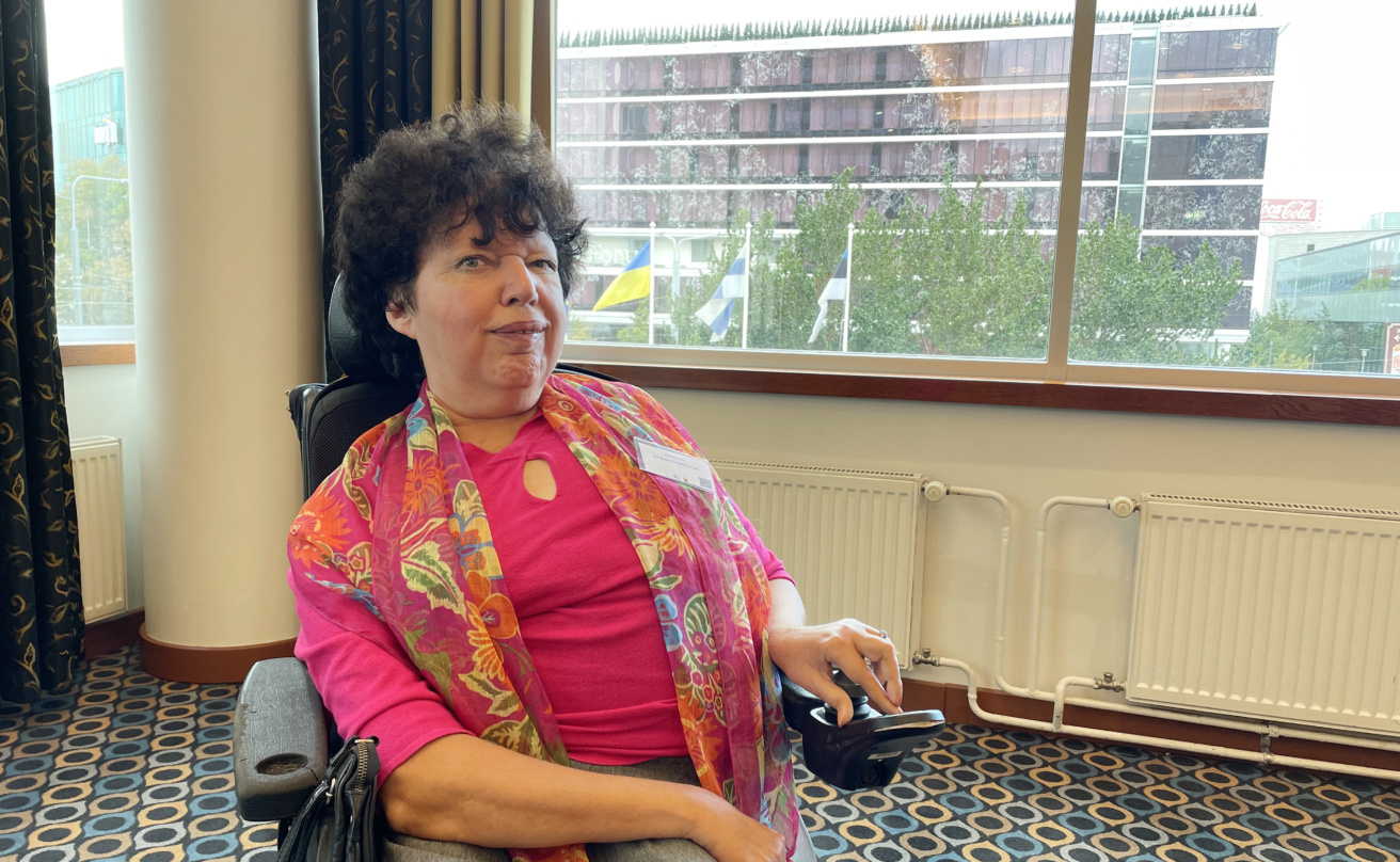 Portrait of Gunta Anca sitting in a wheelchair. Gunta is wearing i bright pink shirt with floral scarf.