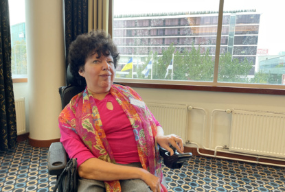 Portrait of Gunta Anca sitting in a wheelchair. Gunta is wearing i bright pink shirt with floral scarf.