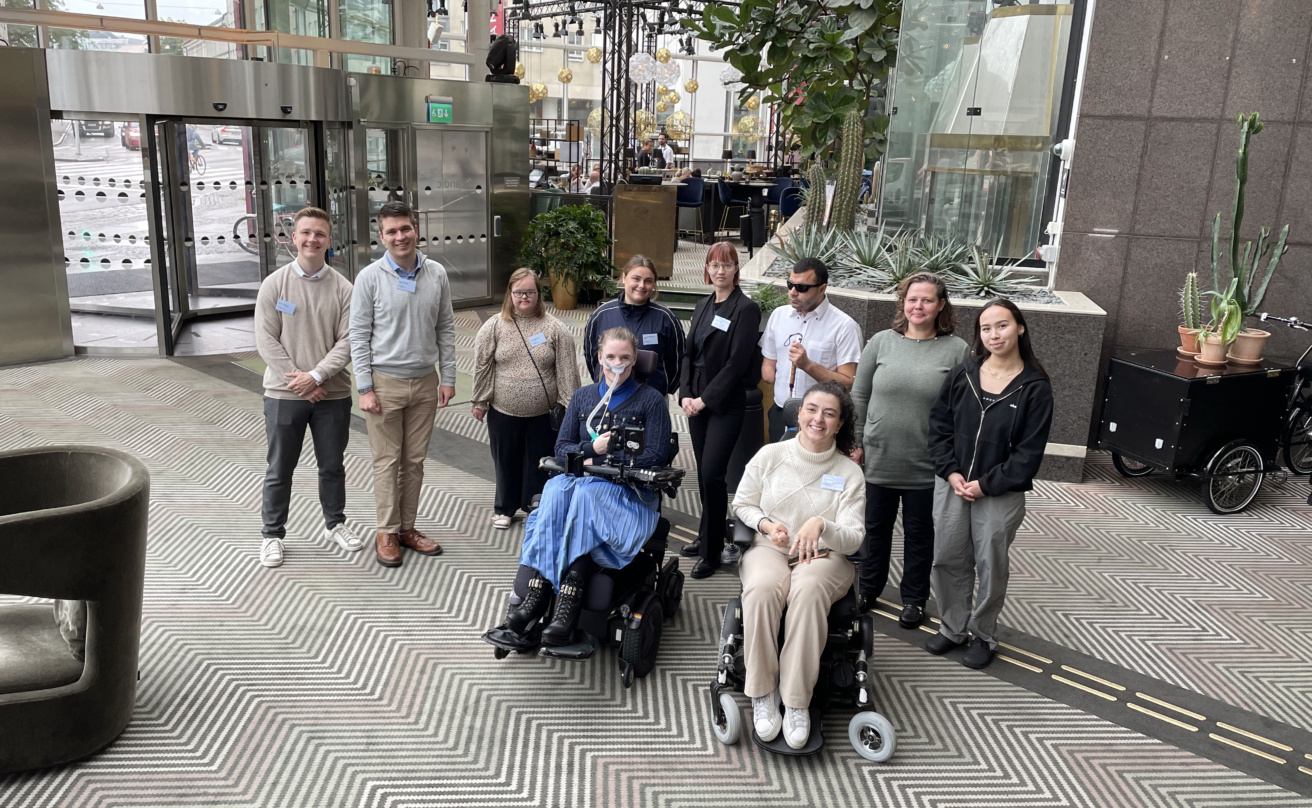 Group picture of ten people in a light hotel lobby. Two are sitting in wheelchairs, eight are standing. Five women, three men. All are young.