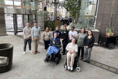 Group picture of ten people in a light hotel lobby. Two are sitting in wheelchairs, eight are standing. Five women, three men. All are young.