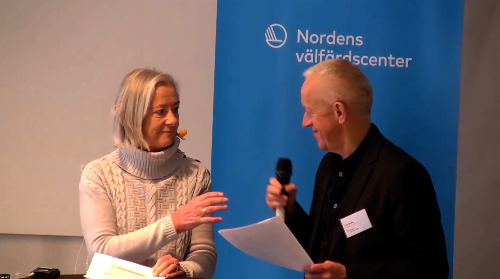 En kvinna övertar mikrofonen från en man. i bakgrunden en blå rollup med texten Nordens välfärdscenter.