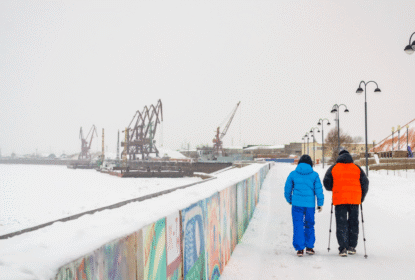 Two people walking with poles along a snowy harbor path, with cranes in the background