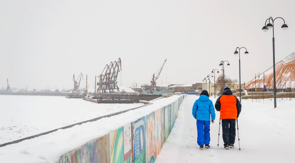 Two people walking with poles along a snowy harbor path, with cranes in the background