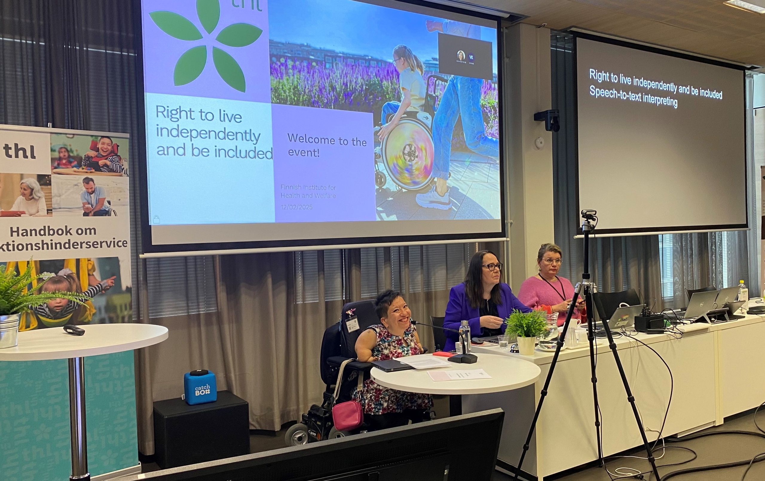 Three women sitting at a desk in a conference room, big screen behind them. One of them, the moderator, is sitting in a wheelchair