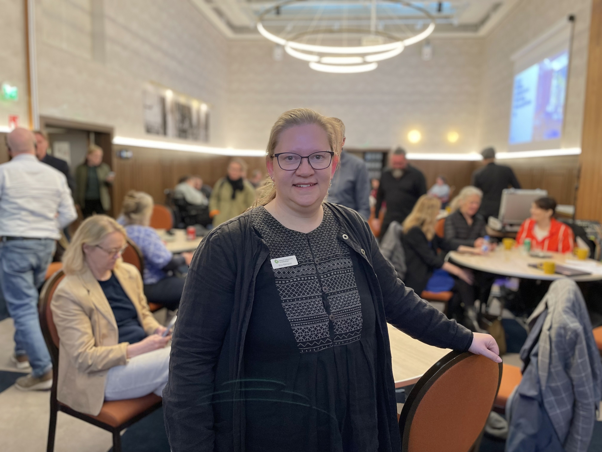 Porträtt av Moa Wahlqvist. Moa har glasögo och mörka kläder. I bakgrunden ett mötesrum med människor som rör sig.