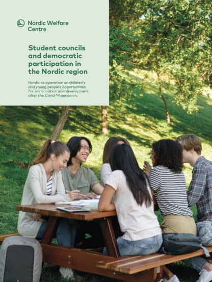 A group of young people sitting around a wooden picnic table in a park, smiling and engaging in discussion or study. The setting is green and sunny, with trees in the background.