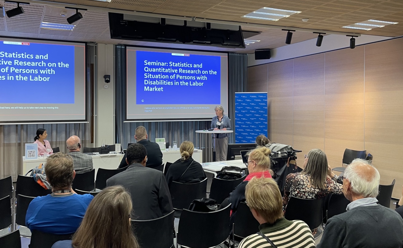 Woman giving a speech in a conference room. Text on powerpoint behind her: Seminar: Statistics and Quantitative Research on the Situation of Persons with Disabilities in the Labor Market