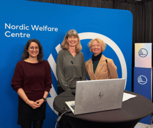 Three women stand smiling behind a round table with a laptop, in front of a blue Nordic Welfare Centre backdrop. Microphones on the table and Nordic Council of Ministers logos in the background. From left to right: Neda Agahi, Lena Dahlberg, and Louise Scheel Thomasen.