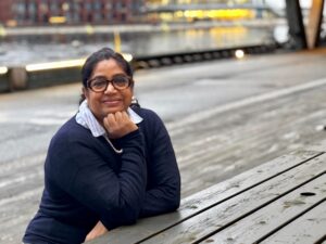 A woman wearing glasses and a dark sweater leans on a wooden table outdoors, with water and illuminated buildings blurred in the background.