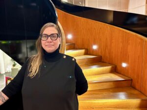 A person stands indoors in front of a curved wooden staircase with black metal detailing, illuminated by small встроen wall lights.