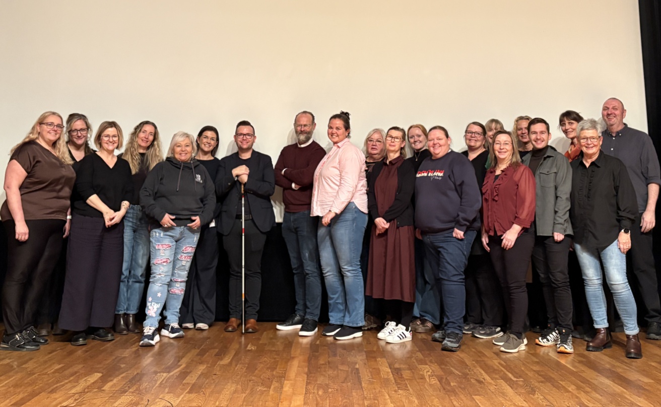 Group photo of seminar participants standing together on a wooden stage in front of a large screen. Around twenty people are posed in a row, smiling at the camera. One participant in the centre is holding a white cane. The group includes professionals from across the Nordic region who took part in the life adjustment seminar.