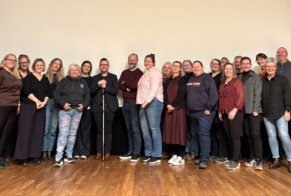 Group photo of seminar participants standing together on a wooden stage in front of a large screen. Around twenty people are posed in a row, smiling at the camera. One participant in the centre is holding a white cane. The group includes professionals from across the Nordic region who took part in the life adjustment seminar.