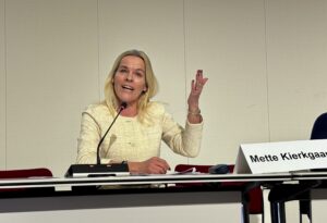 A woman speaks into a microphone while seated at a conference table, gesturing with one hand during a presentation.