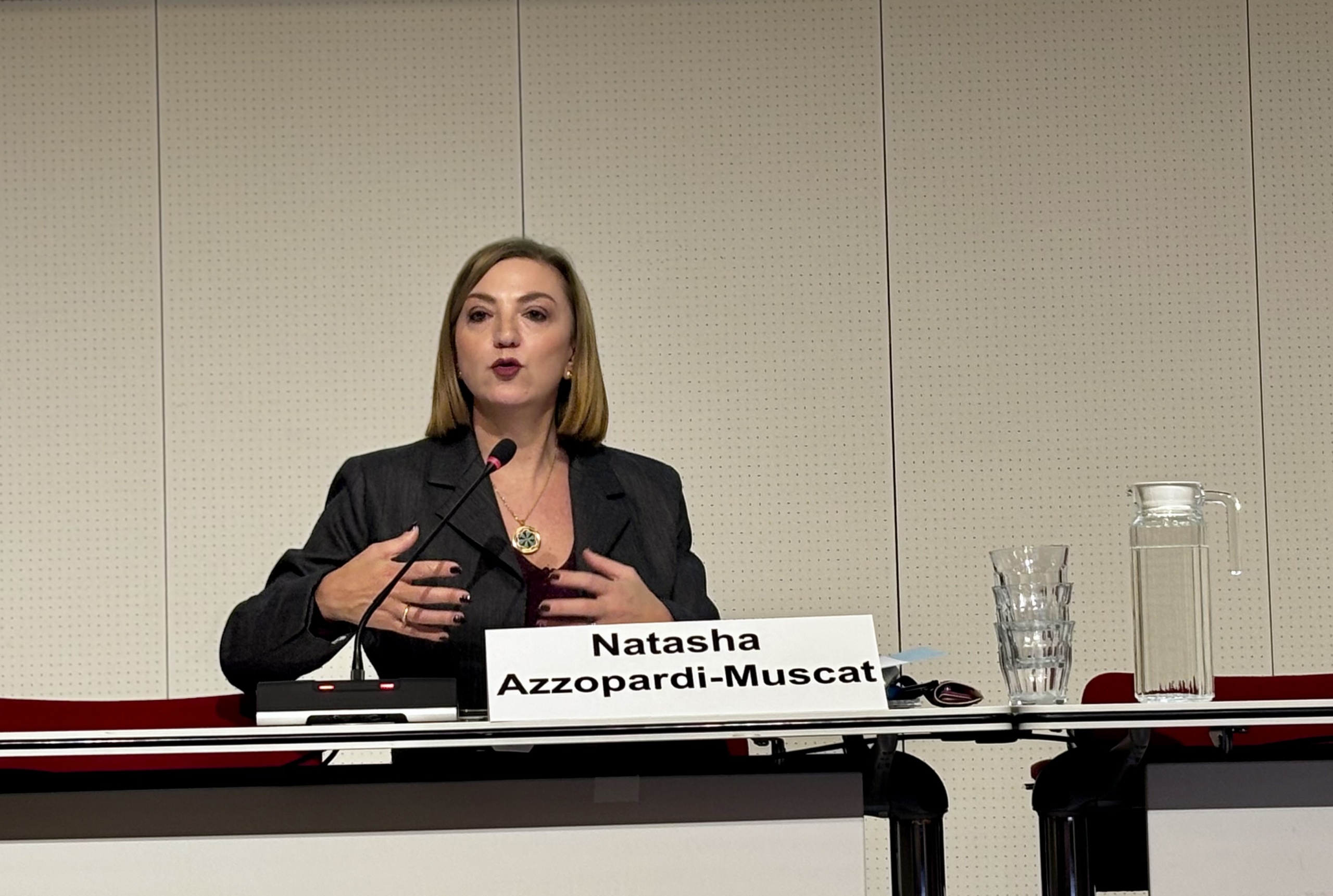 Woman seated at a conference table speaks into a microphone while gesturing with both hands, with a nameplate, water glasses and a water jug visible on the table.