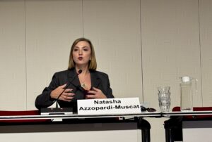 Woman seated at a conference table speaks into a microphone while gesturing with both hands, with a nameplate, water glasses and a water jug visible on the table.