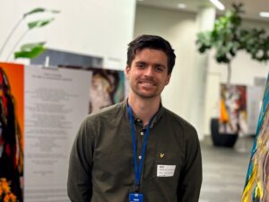 A person stands indoors smiling at the camera, wearing a name badge on a lanyard, with artwork visible in the background.