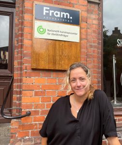 A woman with blond hair, wearing a black blouse, stands smiling in front of a brick building. Behind her is a sign that reads “Fram. Advokatbyrå” and another sign for the “Nationellt kunskapscenter för dövblindfrågor.”