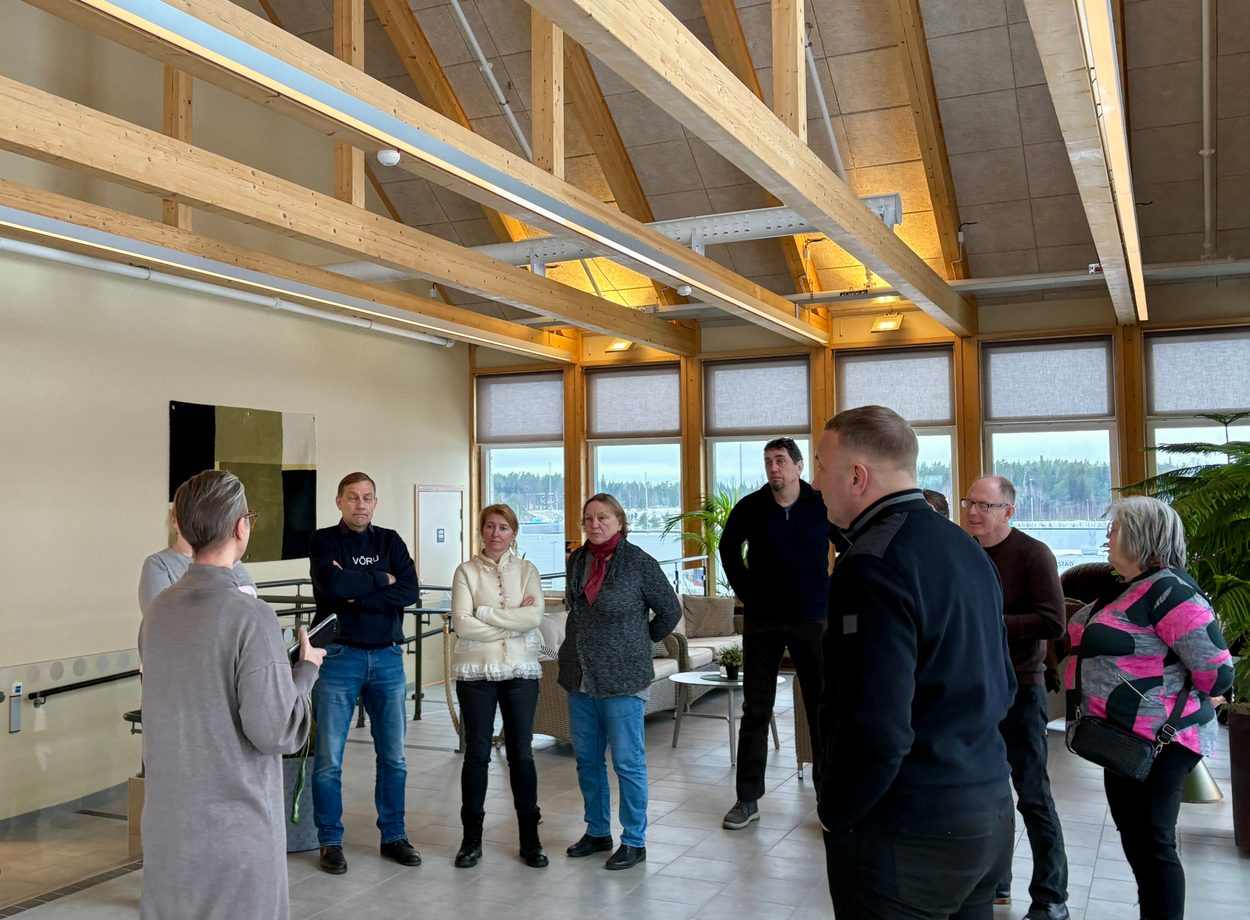 En grupp vuxna står i ett ljust, modernt inomhusutrymme med träbjälkar i taket och stora fönster, och lyssnar på en person som talar till dem. Utanför syns ett snötäckt landskap.