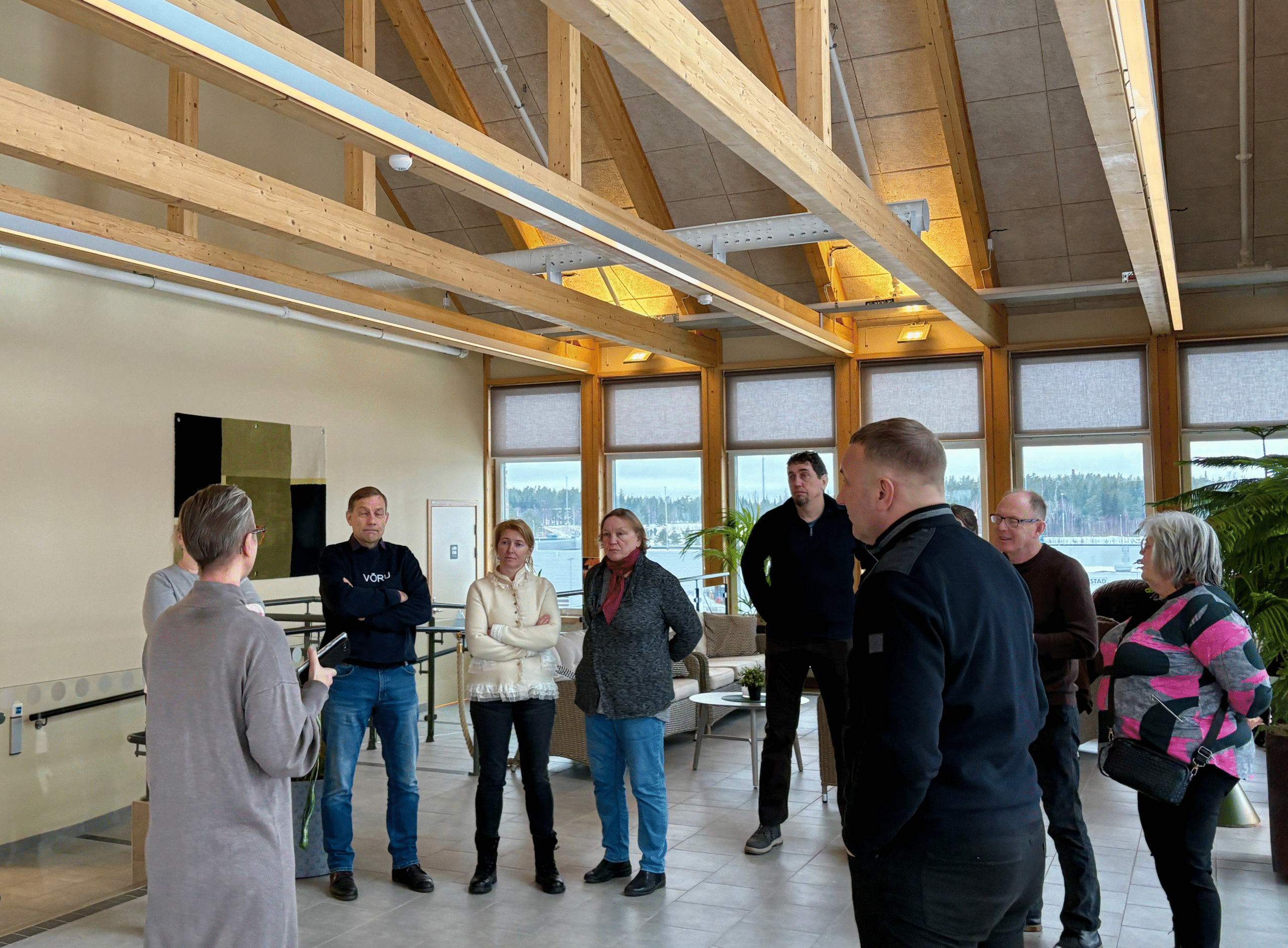 En grupp vuxna står i ett ljust, modernt inomhusutrymme med träbjälkar i taket och stora fönster, och lyssnar på en person som talar till dem. Utanför syns ett snötäckt landskap.